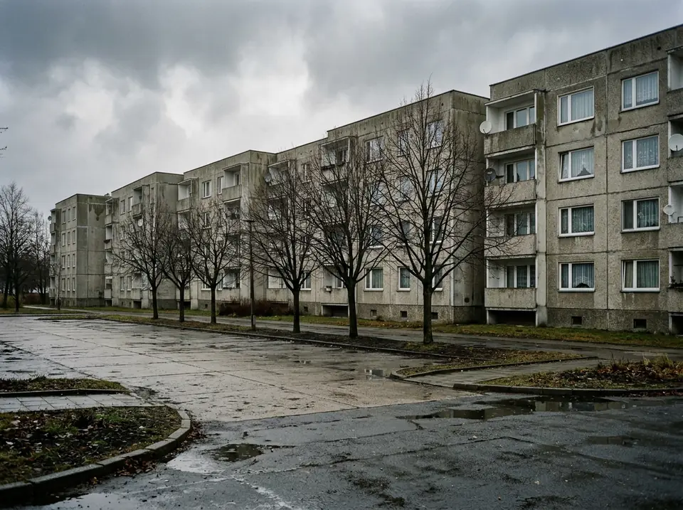 Panelházak sora üres, burkolt parkolófelülettel és csupasz fákkal, borús időben.